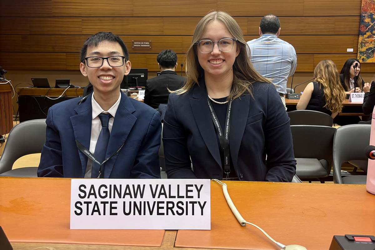 Young man and young woman in dark suits sit at a table with a placard reading Saginaw Valley State University